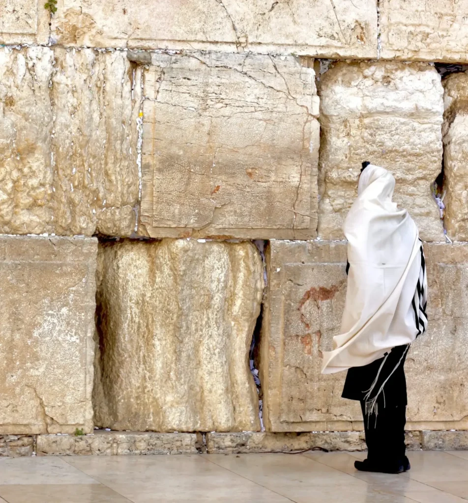 A person wearing a white prayer shawl (tallit) stands in front of the Western Wall (Kotel) in Jerusalem, engaged in prayer. The individual is reciting Kaddish, a Jewish prayer for the deceased. The ancient stone wall is visible in the background.